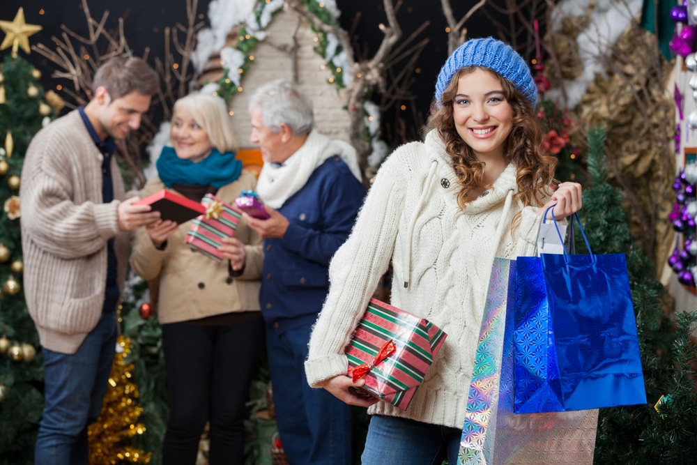 Portrait of beautiful young woman holding Christmas presents and shopping bags with family standing in background at store