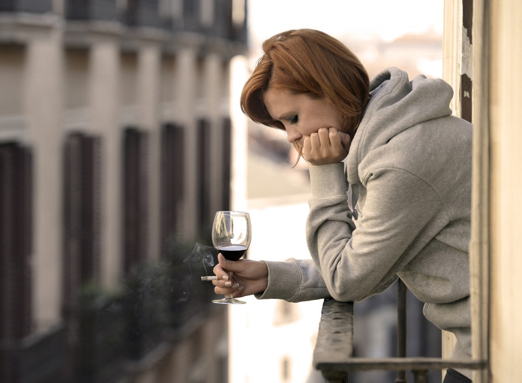 young attractive woman suffering depression and stress smoking drinking glass of wine at the balcony window in pain and grief feeling sad and desperate in urban background